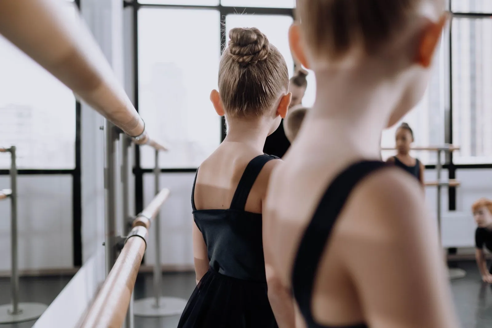 Ballet students in black attire practice in a sunlit studio with mirrors and barre.